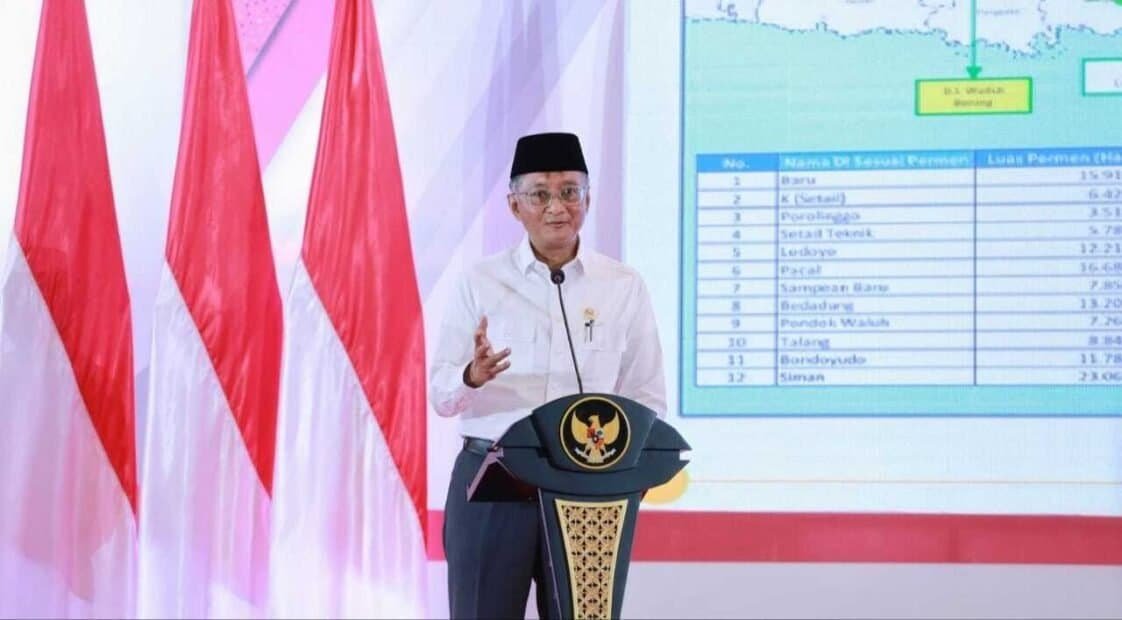 Kiai berbaju putih sedang berbicara di depan podium berlogo Indonesia meninggalkan latar belakang spanduk merah-putih dan layar presentasi.